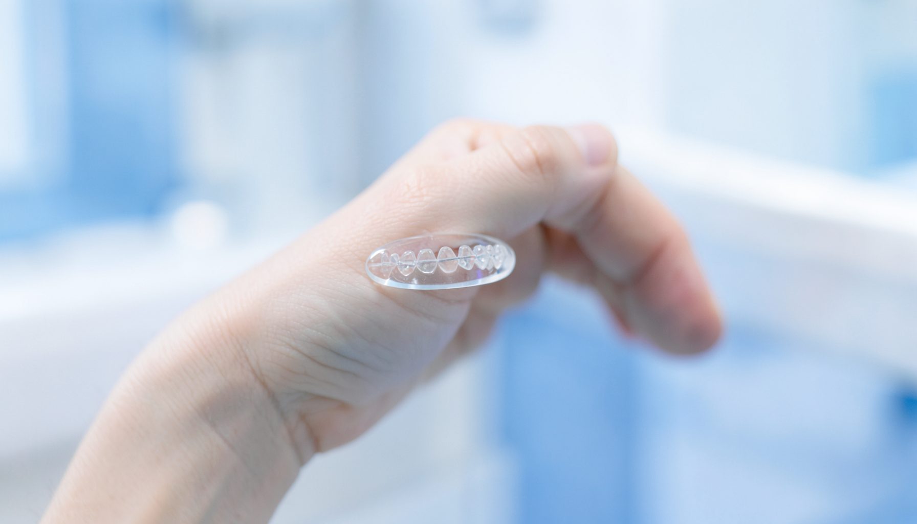 Şeffaf bir Invisalign plak tutan bir elin yakın çekimi; yumuşak mavi ve beyaz tonlarında, temiz ve modern bir klinik atmosferi yansıtan sade bir görüntü.
